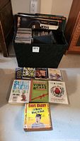 Black basket holding vinyl records and cassette cases, plus several CDs and audio books arranged in front on the floor.