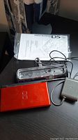 Red Nintendo DS Lite console, clear plastic charger dock, and gray power adapter with cable.