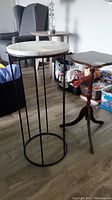 Photo showing the metal plant stand with round marble top and the wooden tripod pedestal plant stand behind it on a hardwood floor with nearby furniture.