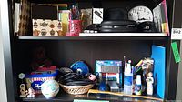 Shelf with various office supplies, wireless headphones in black, Sony Discman portable CD player in silver with a black case, assorted cables coiled in a basket, small decorative items including a small globe, teddy bear, and container with writing utensils.