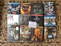 Photo showing 14 action and horror DVD and Blu-ray cases laid out on patterned fabric. Titles clearly visible including Get Out, Ghost Rider, Zodiac, The Exorcist, The Mummy (unopened), Abandon, Captivity, 1408, Asylum, Alone in the Dark, Life (unopened).