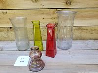 Photo shows five glass vases of varying colors and shapes arranged on a wooden surface.