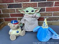 Front view of Baby Yoda plush and plastic toy, ceramic teddy bear bank, and blue first birthday hat