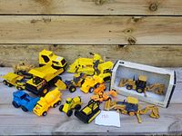 Overview of collection of yellow and blue toy construction vehicles arranged on wooden surface with John Deere backhoe in box visible.