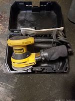 DeWalt random orbit sander placed inside open black carrying case with dust bag attached and power cord coiled.
