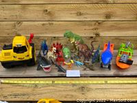 Photo of the lot showing various dinosaur, dragon, and shark figures along with toy boats and a yellow toy bulldozer vehicle on a wooden surface.
