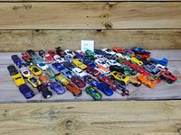 Wide shot showing a total of 70 Hot Wheels toy cars arranged on a wooden floor, showcasing a colorful variety including sports cars, trucks, and fantasy car designs.