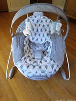 Full view of infant bouncing seat featuring gray and white triangle patterned fabric, mesh sides, padded harness, and overhead toy bar with two plush toys.