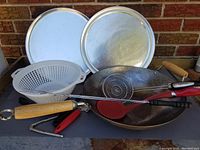Steel wok, two pizza pans, colander, and several utensils grouped together on a surface.