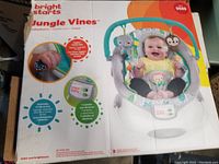Box of Bright Starts Jungle Vines baby bouncer showing front with image of smiling seated baby, toy bar with elephant and sloth plush toys, and features listed including melodies, vibrations, and removable head support.
