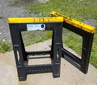 Pair of black plastic folding sawhorses with yellow tops shown open and standing on an outdoor surface.