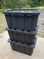 Three black plastic storage bins stacked on top of each other outdoors on concrete with closed lids.
