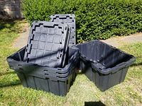 Four black plastic Superbox storage bins and their lids shown stacked outdoors on grass in sunlight.