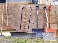 Wide view of four snow shovels and one ice scraper leaning against plywood background showing handle and blade details.