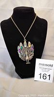 Full view of the metal peacock pendant on a chain displayed on a black stand with auction number card.