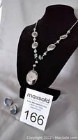 Full view of the necklace displayed on a black bust with black onyx pendant and jade stone beads with matching black onyx stud earrings beside it.