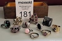 Photo showing 11 medium size costume jewellery rings in various styles and colors arranged on a wooden box with MaxSold auction tag.
