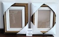Two new wooden picture frames side by side with white protective corner covers still attached, showing gold-tone borders and cream color panels.