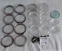 Photo showing large clear glass lids and smaller clear glass lids along with metal clips for Mason jars, all laid out on cloth surface for detail and condition.