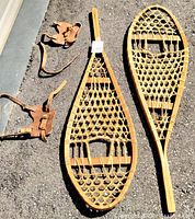 Two wooden snowshoes with rawhide webbing and two leather harnesses laid on pavement in sunlight showing overall lot contents.