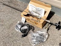 Full view of the BBQ rotisserie components including the rotisserie rod, electric motor with cord, skewers, and assembly instructions in original box.