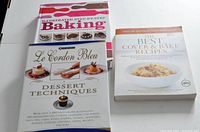 Top view of three hardcover baking books titled 'Illustrated Step-by-Step Baking,' 'Le Cordon Bleu Dessert Techniques,' and 'The Best Cover & Bake Recipes'.
