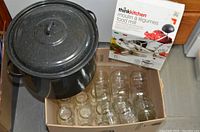 View of large black speckled enamel canning pot with lid beside a cardboard box containing clear glass canning jars and a ThinkKitchen food mill in box.
