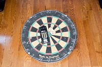 Full view of the dart board on a wood floor, circular with numbers around edge, 'Made in England' printed at the bottom.