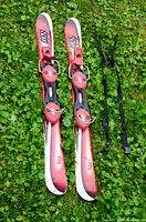 Red and pink Elan kid's skis with bindings placed on green grass alongside adjustable black ski poles.