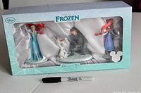 Front box photo showing four Frozen character ornaments secured inside a display box with a clear window, including Elsa, Olaf, Kristoff, and Anna, along with a Sharpie marker for scale.