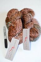 Two skeins of yarn with visible Hand Maiden tags showing the 'Maiden Hair' series and blend of silk kid mohair and nylon. The yarn is multicolored with soft gradient earth tones and a slightly fuzzy texture.