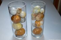 17 various natural rock and mineral eggs displayed in two clear cylindrical glass holders on a white surface, showing a range of colors from white, cream, yellow, brown, and some translucency.