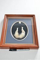 Front view of framed sculpted fur artwork showing two white wolves sitting and howling at a moon within an oval matting and wooden frame.