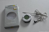 White circular digital thermometer with LCD screen and arrow buttons, connected to metal temperature probe by wire, displayed alongside original ThermoWorks DOT box with noticeable wear.