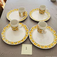 Set of four ceramic cups sitting on matching plates with yellow floral pattern around rims.
