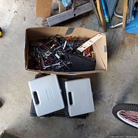 Photo showing a cardboard box filled with assorted hand tools and accessories on the ground, alongside two closed gray plastic tool carrying cases.