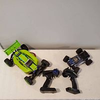 Two off-road style remote control cars with their respective handheld remote controllers placed on a white surface.