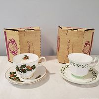 Two Royal Crest bone china tea cups and saucers displayed with their original boxes in the background. One cup set features holly and pine cone motif, the other a green leaf pattern.