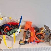 Lot of Hot Wheels track pieces and accessories inside a patterned tote bag and arranged on table including orange and gray curved track sections, and some toy parts related to dinosaur theme.