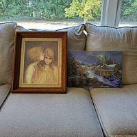 Framed print of two children by M. Kane on left, light-up winter village scene on right, both displayed on a couch.