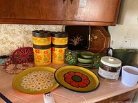 Four large floral-patterned 1970s canisters, two floral plates, two crocheted pot holders, a crocheted hotpad and a vintage stainless steel coffee mug arranged on a tile countertop with floral wallpaper background.