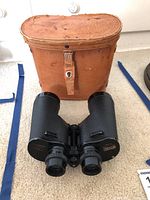 Front view of binoculars placed in front of the brown leather case on carpeted floor.