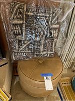 Brown heart-shaped ottoman with attached label and large square patterned pillow wrapped in plastic, arranged together in a room corner.