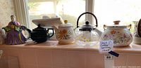 Wide view of multiple ceramic and glass teapots arranged on a tiled surface, including floral designs and a figurine teapot.
