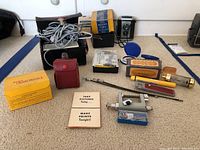 Photo showing multiple vintage photography items including Brownie Reflex camera, flashes, cables, cases and small accessories laid out on floor.