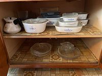 View of shelf with multiple Corelle and Pyrex baking dishes and bowls, clear lids, and serving ware.