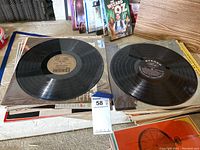 Top view of two vinyl records laid flat, showing labels and part of collection in background.