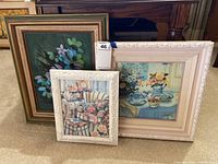 Three framed paintings displayed leaning against wood furniture: floral still life with tea set, floral bouquet on dark green background signed K. Adams, and interior floral chair scene.