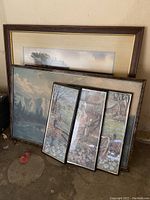 Five framed landscape artworks leaning against a wall on a concrete floor, showing varying sizes and styles of nature images.