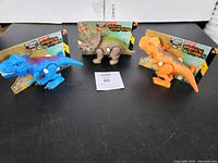 Set of three packaged new wind-up dinosaur toys on a black surface: blue T-Rex, Triceratops, orange T-Rex.